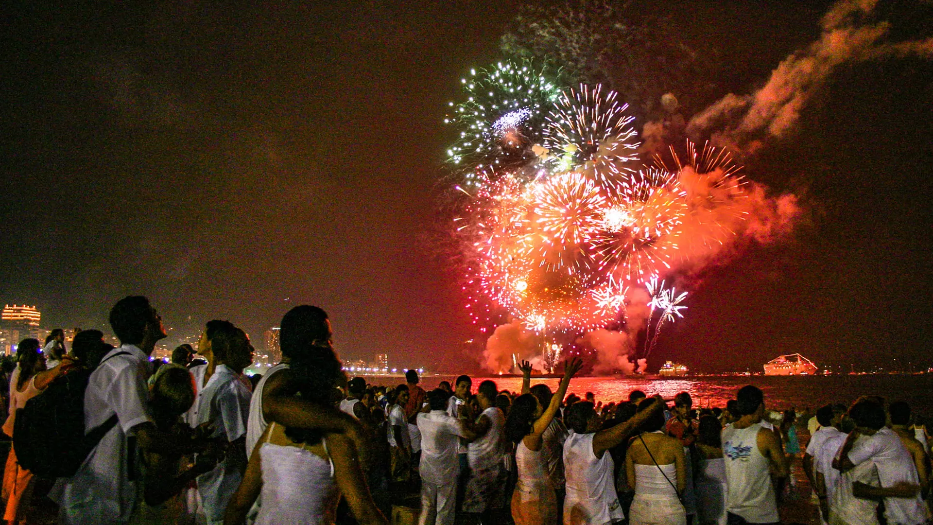 Multidão reunida na orla assistindo shows de fogos de artifício iluminando o céu noturno à beira-mar.